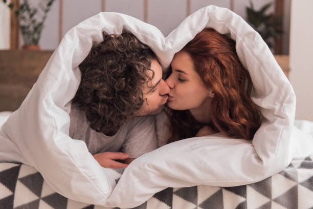 beautiful couple is kissing while lying in bed wrapped in blanket